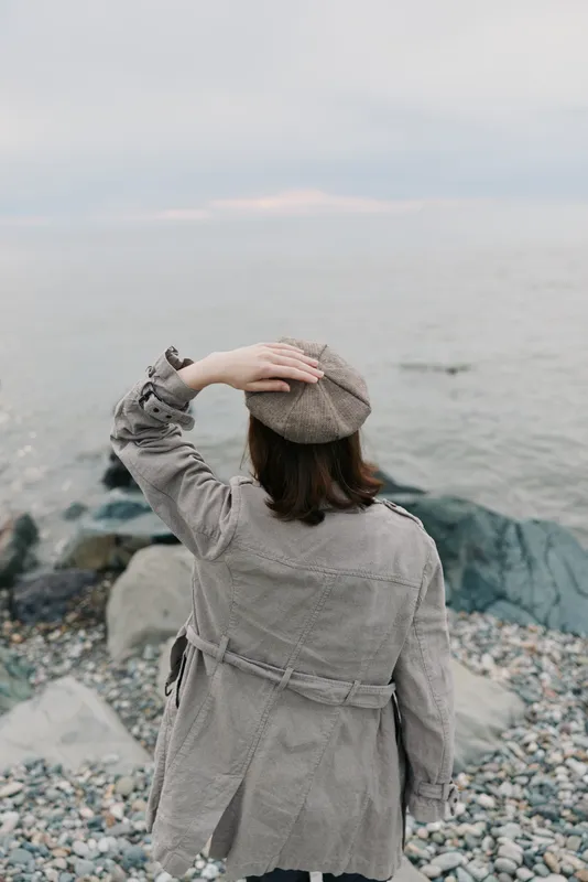 Woman wearing a coat and knitted beret stands on rocky shore facing calm sea. Overcast sky and pebbles create a peaceful, contemplative atmosphere by the water.
