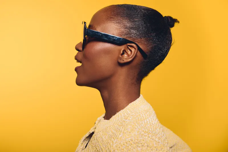 Profile of a dark skinned African American woman wearing black sunglasses and a beige sweater against a vibrant yellow background. Concept of fashion, style, and confidence.
