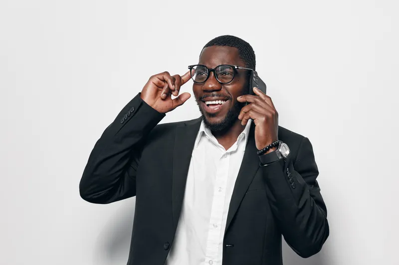 African American man with dark skin talking on phone and smiling while adjusting glasses in casual business attire on white background. Positive communication concept.