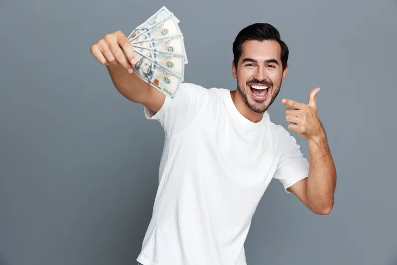 Happy smiling man holding cash bills, looking at the camera with excitement, dressed casually in a white t-shirt against a plain, neutral background.