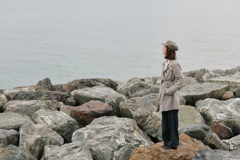 Young woman in warm clothing stands on rocky shore looking at calm sea, wearing hat and coat. Concept of solitude, nature, and cold weather outdoors by the water.