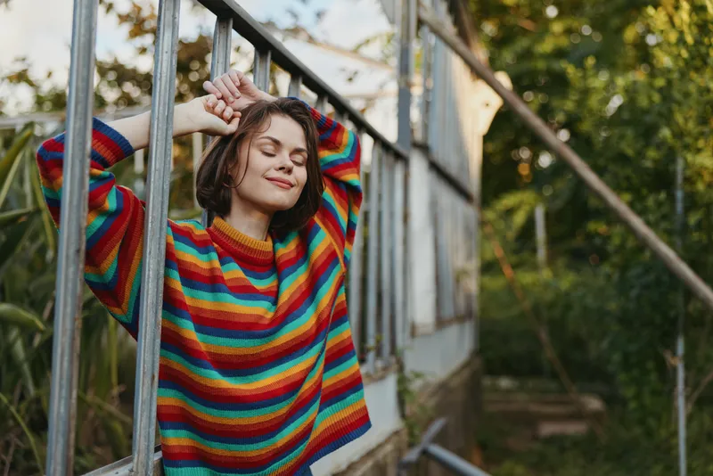 Woman in a rainbow sweater relaxing with eyes closed leaning on greenhouse window, portrait of a calm young adult enjoying nature, warm sunlight and peaceful outdoors moment.