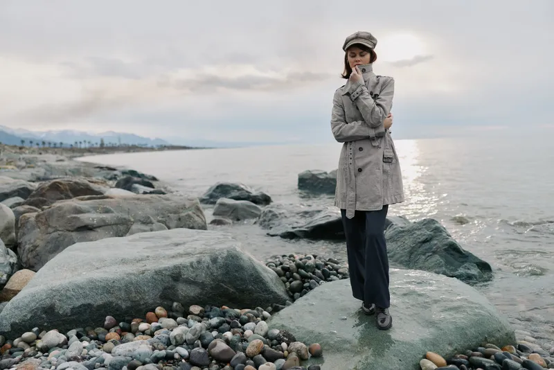 A thoughtful woman in a grey coat and hat stands on rocky shore near calm sea under cloudy sky. Concept of autumn fashion, contemplation, and nature by the water.