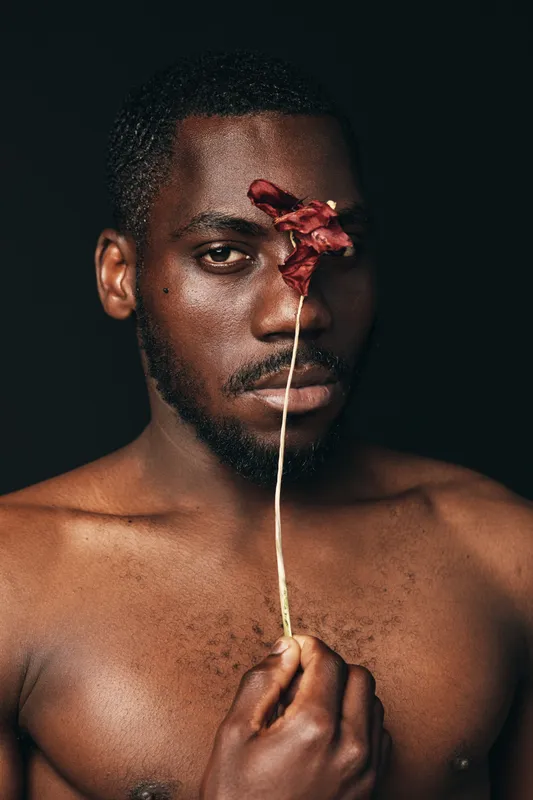 African man with dark skin holding dried flower in front of face in creative conceptual visual style. Portrait with bare chest and artistic expression on black background.
