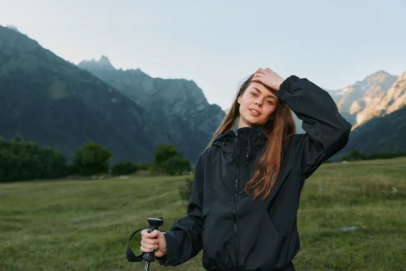 A young woman stands in a mountainous landscape, shading her eyes with one hand and gripping a trekking pole with the other. She wears a dark windbreaker, ready for outdoor exploration and discovery