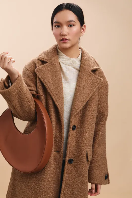 Asian woman wearing a tan coat and light knit sweater stands against a neutral backdrop, carrying a curved brown handbag and looking calm yet confident.