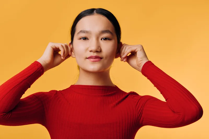 Young woman in red sweater adjusting hair against orange background, looking at camera with calm expression, studio portrait of female model with natural makeup.