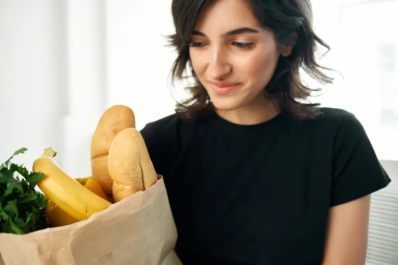Женщина в черной футболке держит пакет с продуктами: овощами и фруктами, символизирующими здоровое питание