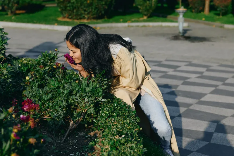 Woman in a beige coat leans toward blooming flowers in a sunlit garden, smelling the fragrance and enjoying a moment of outdoor nature discovery among lush greenery