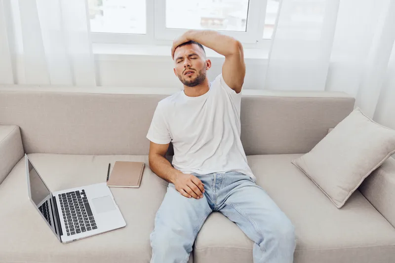 Frustrated man sitting on a sofa with a laptop, reflecting modern work challenges. Feeling overwhelmed in a bright, minimalist home office atmosphere.