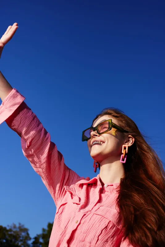 Happy young woman in oversized sunglasses waving against a clear blue sky, wearing a pink shirt, showcasing carefree summer vibes and vibrant emotions