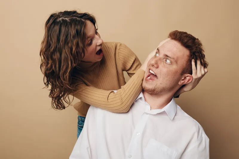 Surprised woman with curly hair grabs shocked man by hair in beige sweater and white shirt. Emotional interaction between young adults on neutral background. Concept of conflict and reaction.