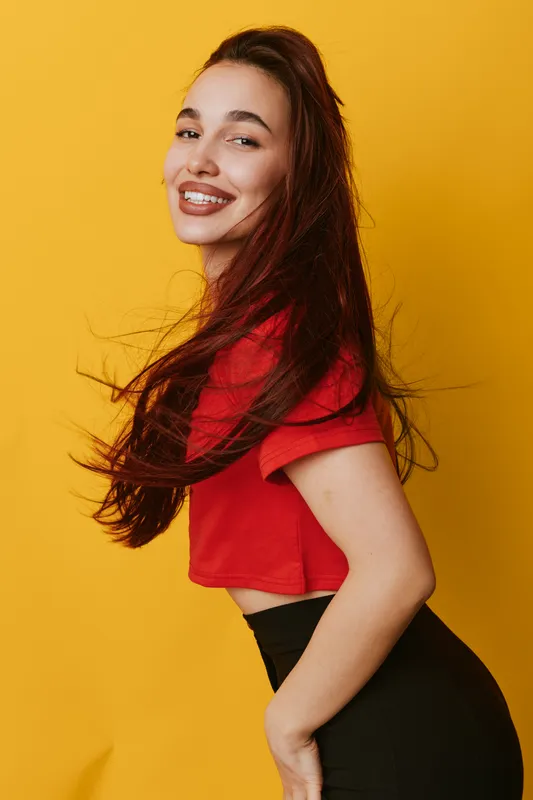 Young woman smiling with long hair wearing red crop top and black pants standing against yellow background. Portrait of happy female with casual style and confident pose.