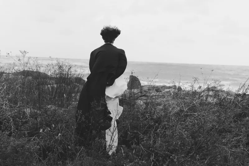 Coast shoreline silhouette with a back view of a figure in a long coat facing the ocean, moody horizon and wild grasses creating a calm, contemplative scene