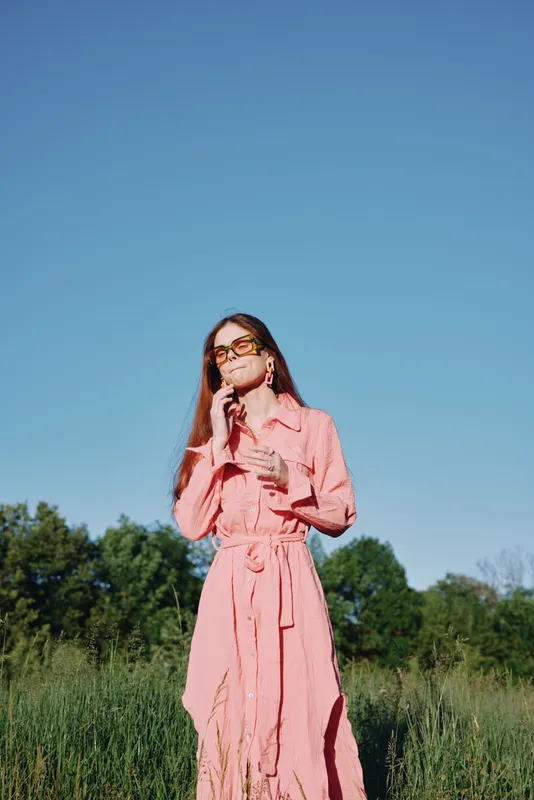Stylish woman in a pink dress expressing confidence in a lush green field under a clear blue sky, embodying beauty and tranquility in nature