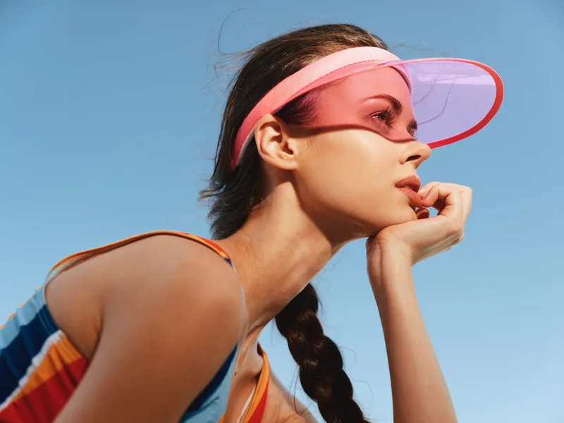 Portrait of a young athletic woman in a pink visor, braided hair, and colorful tank top, gazing toward the distance against a clear blue sky.