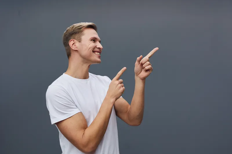 Smiling man in casual white t-shirt pointing with both hands right side, cheerful expression, studio portrait with plain gray background, positive mood, human emotion
