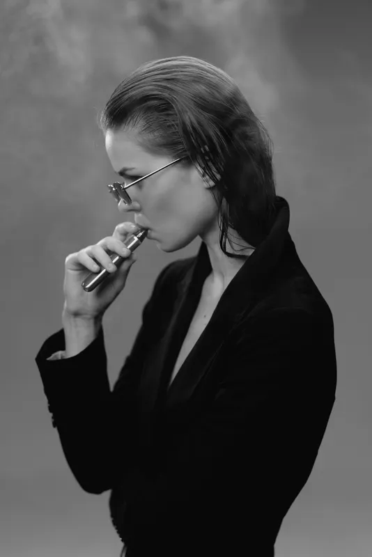 Black and white portrait of a pensive woman vaping with glasses in a dark blazer.