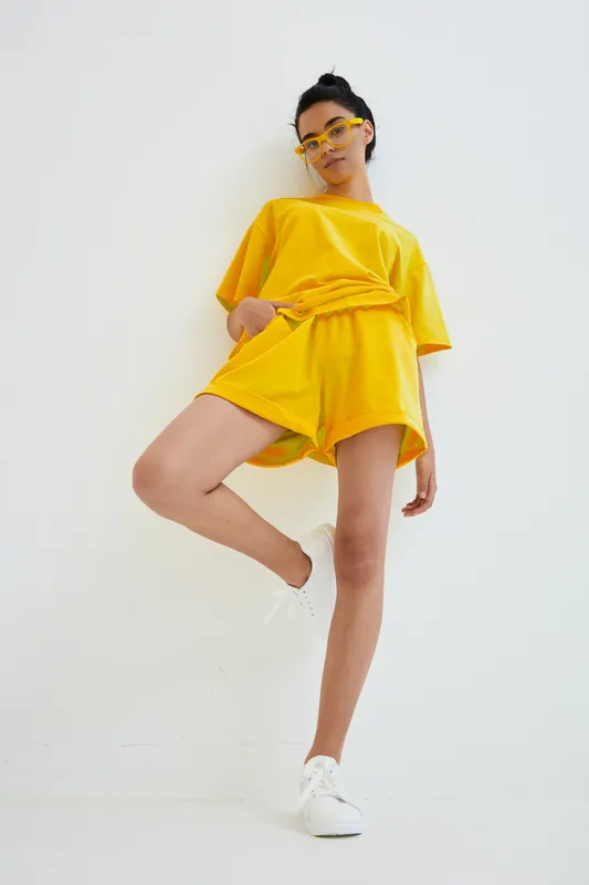 Young woman wearing bright yellow casual outfit and white sneakers poses confidently against white wall, showcasing summer fashion style with sunglasses and relaxed mood.