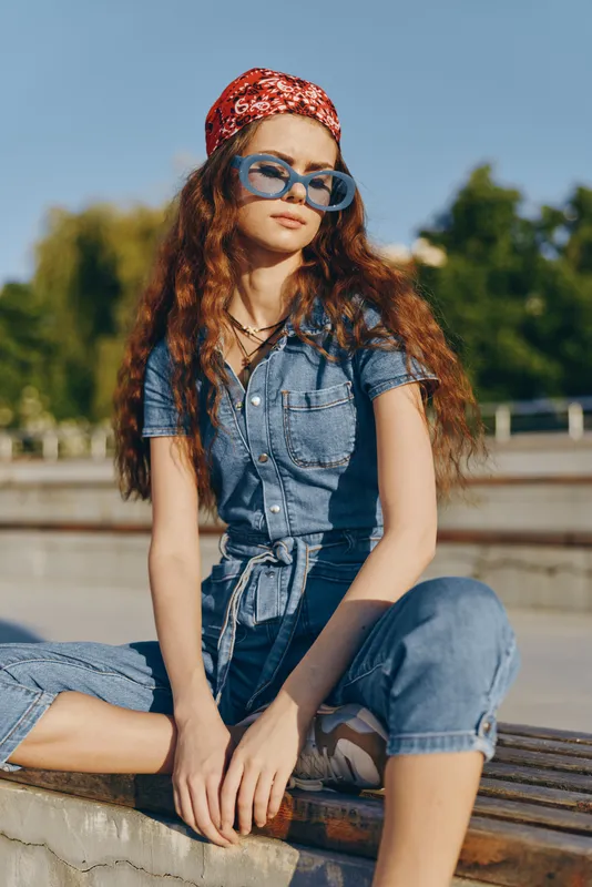 Lifestyle woman in modern boho-western denim street style with red bandana and blue sunglasses, outdoors with natural light and film color effect, relaxed and stylish pose.
