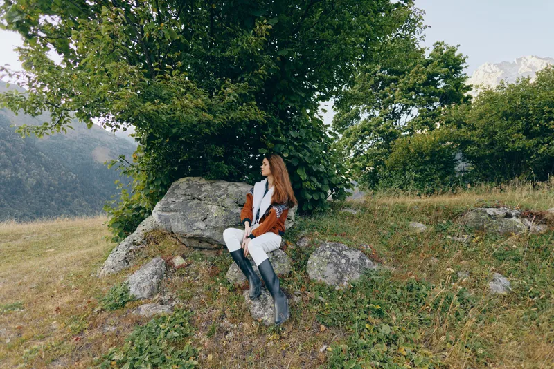 Woman sitting on rock under tree in mountain meadow outdoors, portrait of a traveler in boots and warm coat enjoying the landscape and peaceful autumn nature with distant peaks.