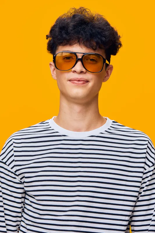 young man wearing striped shirt and large yellow sunglasses poses with slight smile on vibrant orange background, casual style and curly hair visible