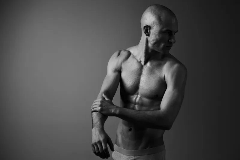 Black and white image of a man showcasing his muscular physique with a contemplative expression, highlighting strength and emotion in a minimalist setting.