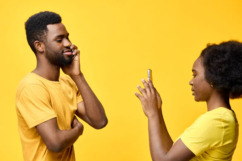 A cheerful Black couple engaging in a fun phone conversation against a vibrant yellow background, showcasing joy and connection with warm tones