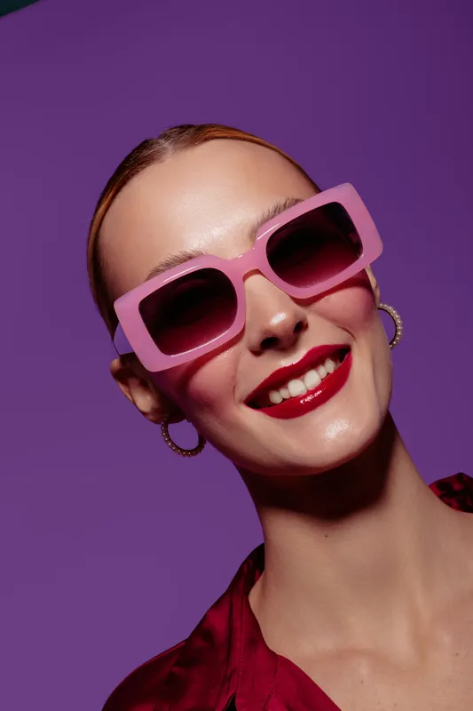 Smiling woman with pink sunglasses and red lipstick poses against purple background. Fashion portrait with hoop earrings and sleek hairstyle. Concept of style and confidence.