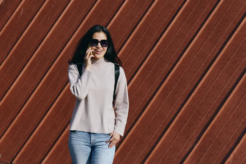 Woman in a light sweater and jeans with sunglasses and a backpack stands against a diagonal wooden wall, showcasing casual fashion and confident outdoor style.
