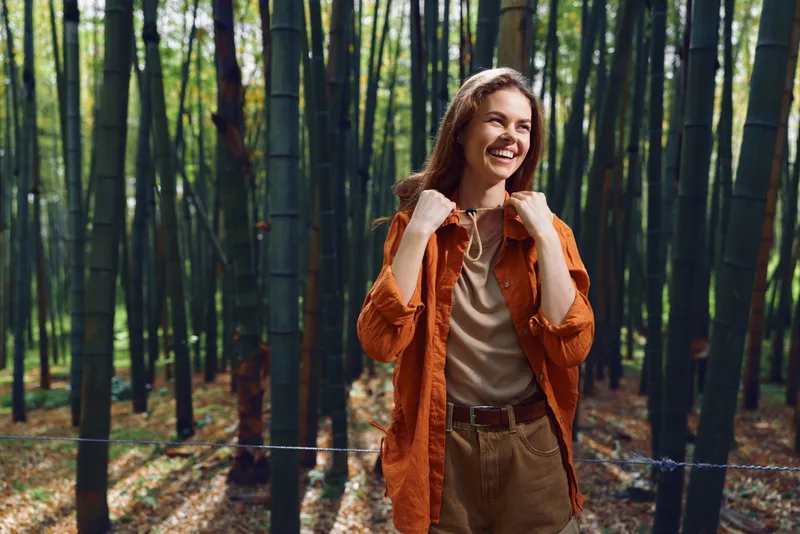 Woman smile bamboo forest nature jacket outdoor happy portrait of a traveler walking along a green grove path, wearing orange jacket and casual outfit, enjoying sunlight and tranquility.