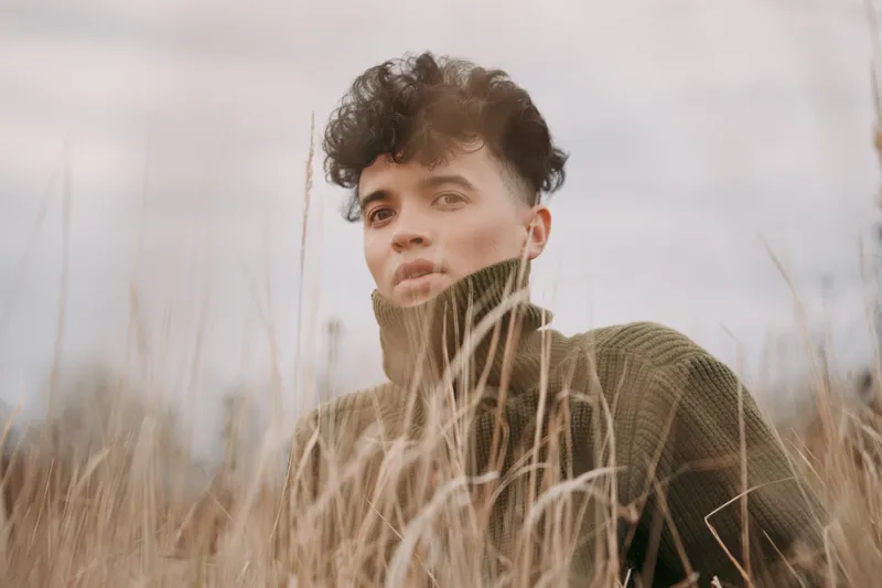 A young person in a textured sweater peers through tall dry grass, offering an authentic and credible portrait of quiet introspection in soft natural light and muted earthy tones.