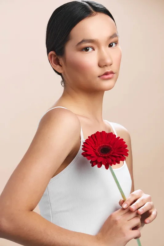asian woman smiles softly as she holds a red flower against a neutral backdrop, presenting elegance and calm in a studio portrait with subtle light and gentle mood.