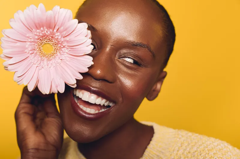 Happy dark skinned African woman smiles holding pink flower near face on yellow background. Concept of natural beauty, happiness, skincare, and floral wellness with bright studio lighting.