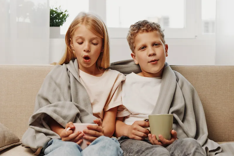 Two surprised children wrapped in gray blankets sit on a sofa holding cups in a bright cozy room. Concept of childhood, warmth, comfort, and home relaxation.