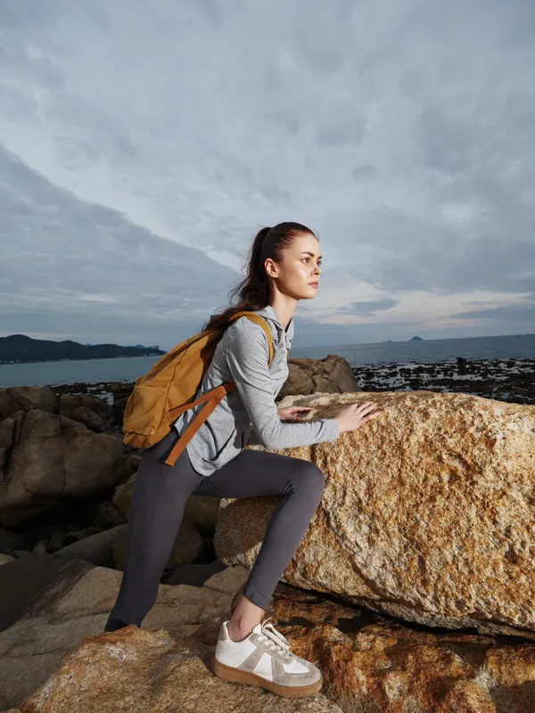 Young woman with backpack climbing on rocks by the sea, showcasing determination and adventure in a sporty outfit under a cloudy sky
