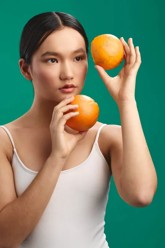 Asian woman holds two oranges and studies them against a vivid backdrop. The composition highlights bright color, calm focus, and a playful still life mood.
