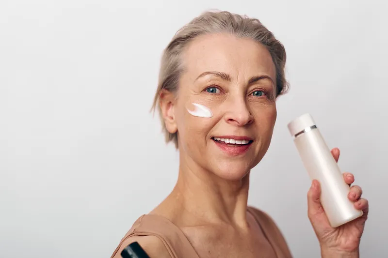 Smiling middle aged woman applying moisturizing cream on cheek holds white cosmetic bottle closeup. Concept of skincare routine, anti aging, facial care, beauty treatment, and healthy skin.