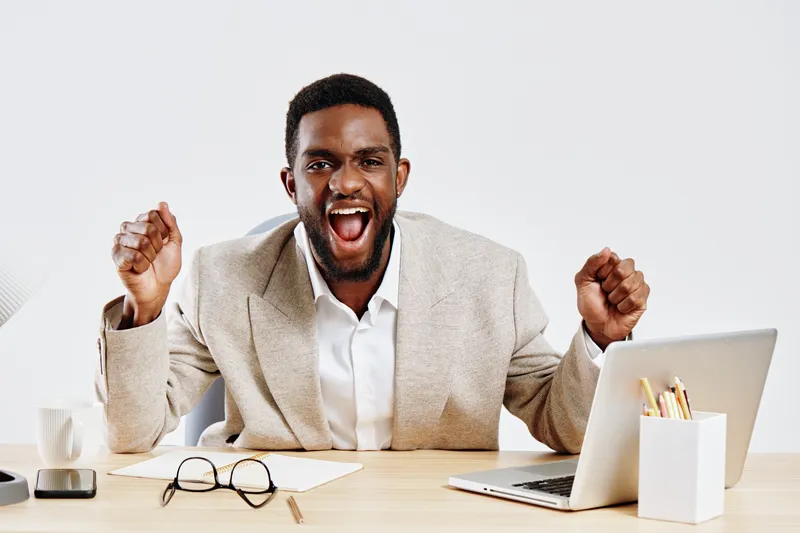Confident happy young African American man in beige blazer celebrating success at work, clenched fists, smiling broadly, sitting at desk with laptop, glasses, and office supplies. Office lifestyle con