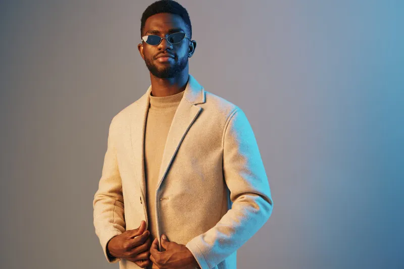 Stylish African man in a beige coat and sunglasses against a blue background, exuding confidence and modern fashion