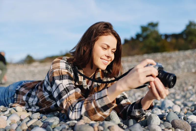 A woman photographer lying on a pebble beach, holding a camera and capturing a candid moment. Sunny outdoor scene with casual clothing, relaxed mood, and natural landscape.