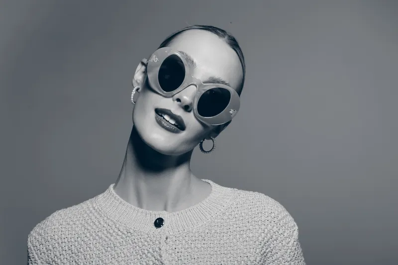 Portrait of a young Caucasian woman with pale skin wearing round sunglasses and a knitted sweater. Studio portrait with neutral grey background.