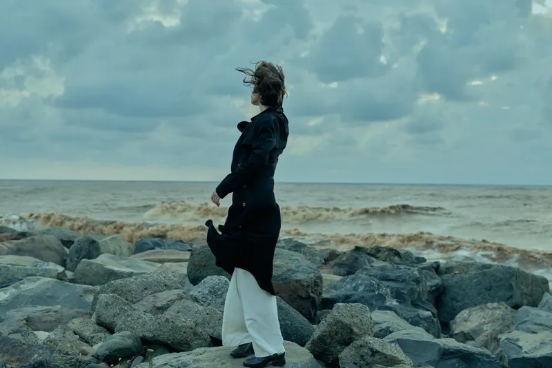 Windy coastal scene with a woman standing on rugged rocks, hair and coat billowing, watching the sea under a moody sky, dramatic natural landscape and calm horizon