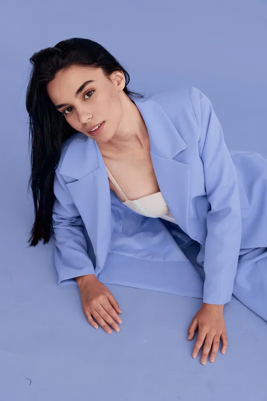 Young woman in blue suit lying on pastel background with relaxed and confident expression, showcasing modern fashion and natural beauty in soft lighting.