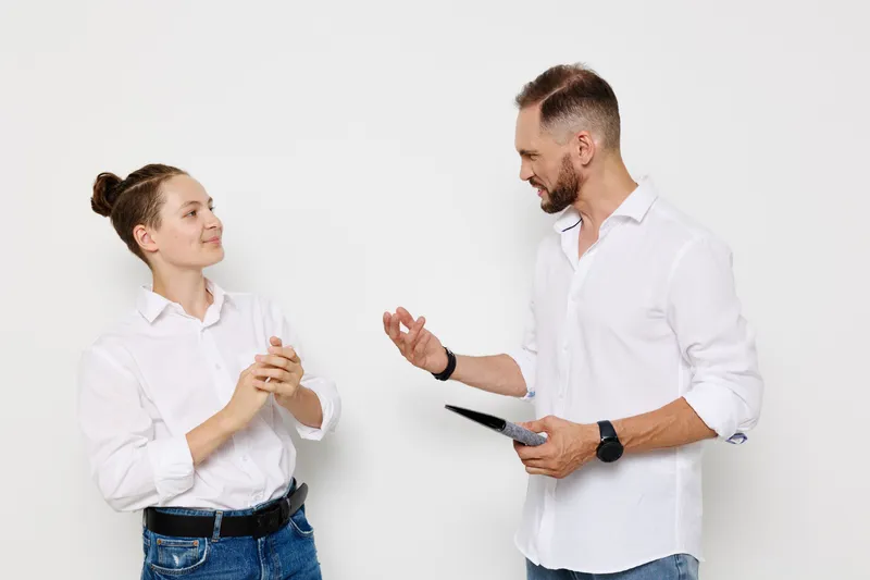 A professional man and woman in white shirts stand against a neutral backdrop, sharing a friendly moment as they discuss ideas, showing confidence, collaboration, and clear communication.