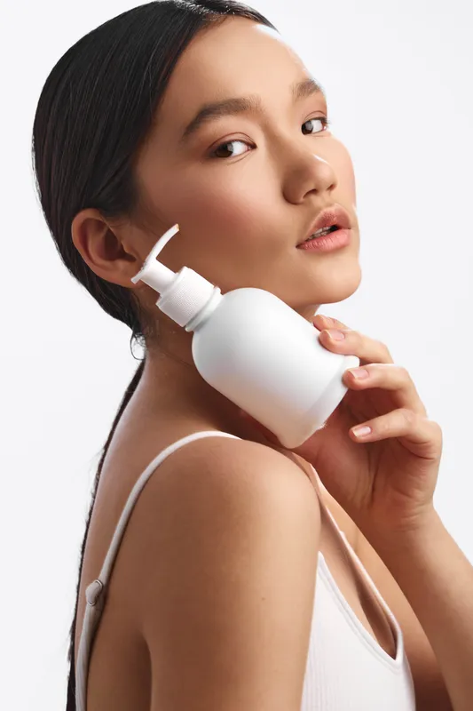 asian woman holds a pump bottle near her face in a bright studio, showcasing skincare product use. she exudes calm confidence, creating a clean, modern beauty moment.