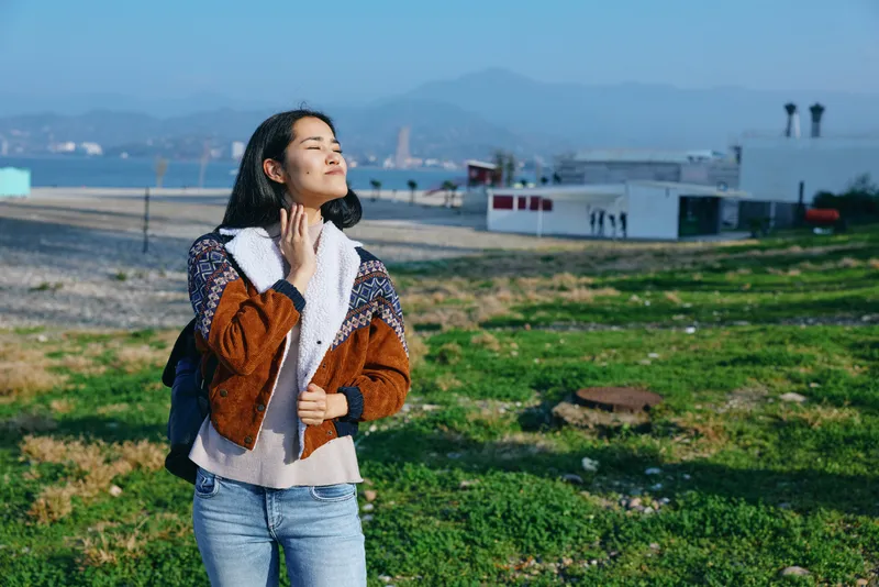 Active, stylish woman enjoying outdoor breeze on a sunny day, wearing a brown and white shearling jacket, jeans, standing on a green field near a coastal town with modern buildings