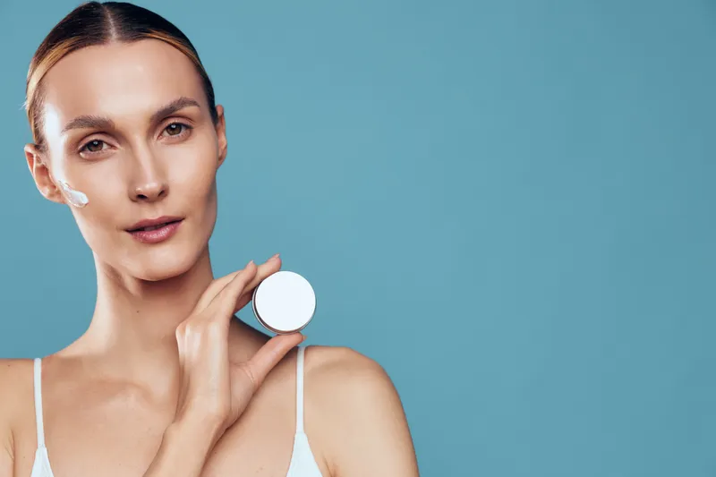 Woman skincare beauty portrait, applying moisturizer with cotton on face, spa routine, smooth skin and natural glow, close up, blue background, soft studio light, minimal copy space, calm