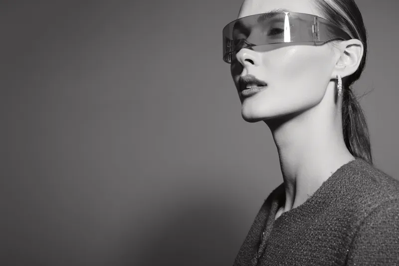 Elegant white woman with pale skin wears futuristic glasses and earrings, posing in black and white studio. Concept of fashion and modern style.