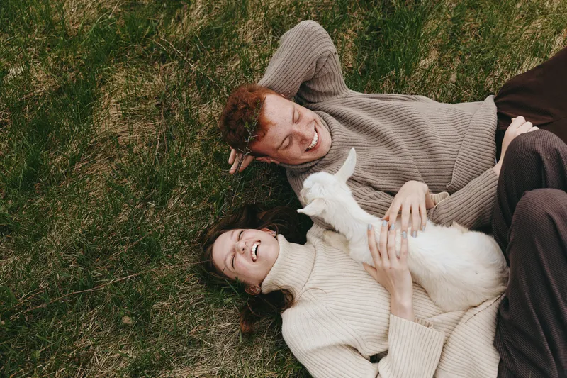 Young couple lying on grass with a white baby goat, enjoying a relaxed moment together in casual sweaters with joyful expressions in a natural outdoor setting.